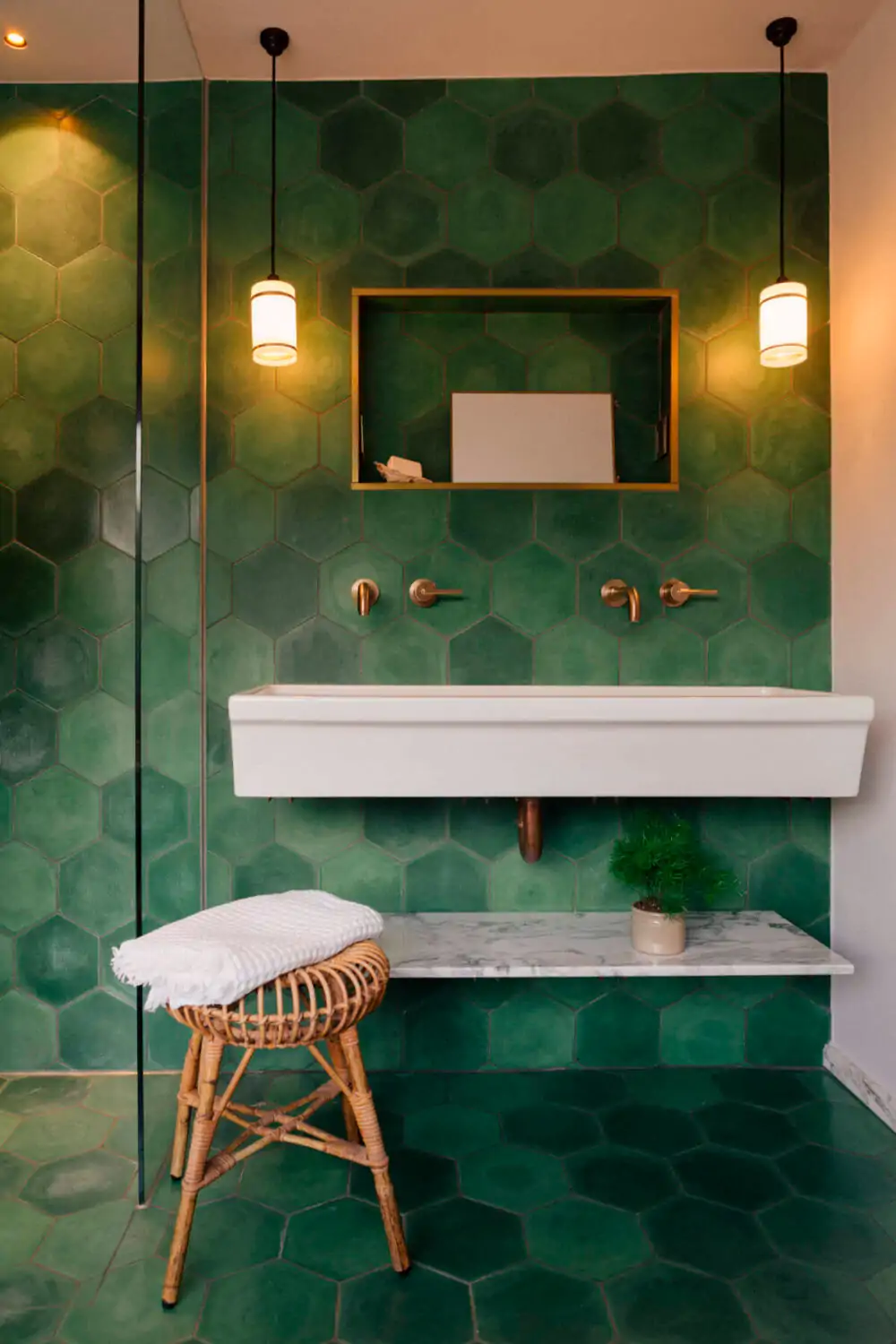 Baño con azulejos hexagonales verdes, lavabo suspendido blanco, grifería dorada y detalles naturales en mármol y ratán.