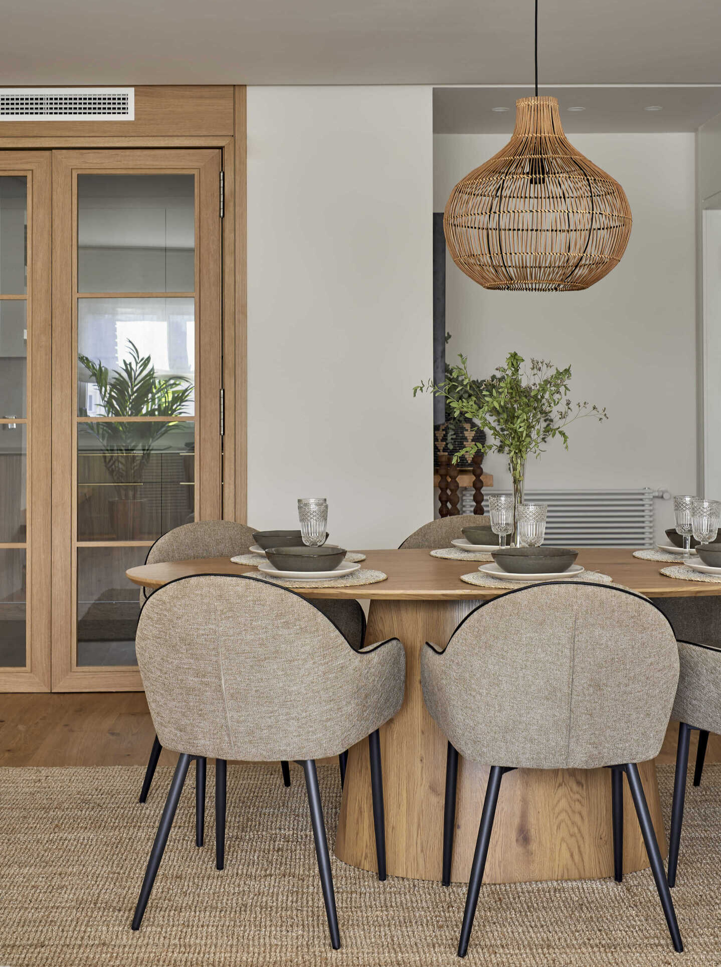 Comedor con mesa de madera, sillas tapizadas en tonos beige, lámpara colgante de fibras naturales y vitrinas de madera con puertas de cristal.