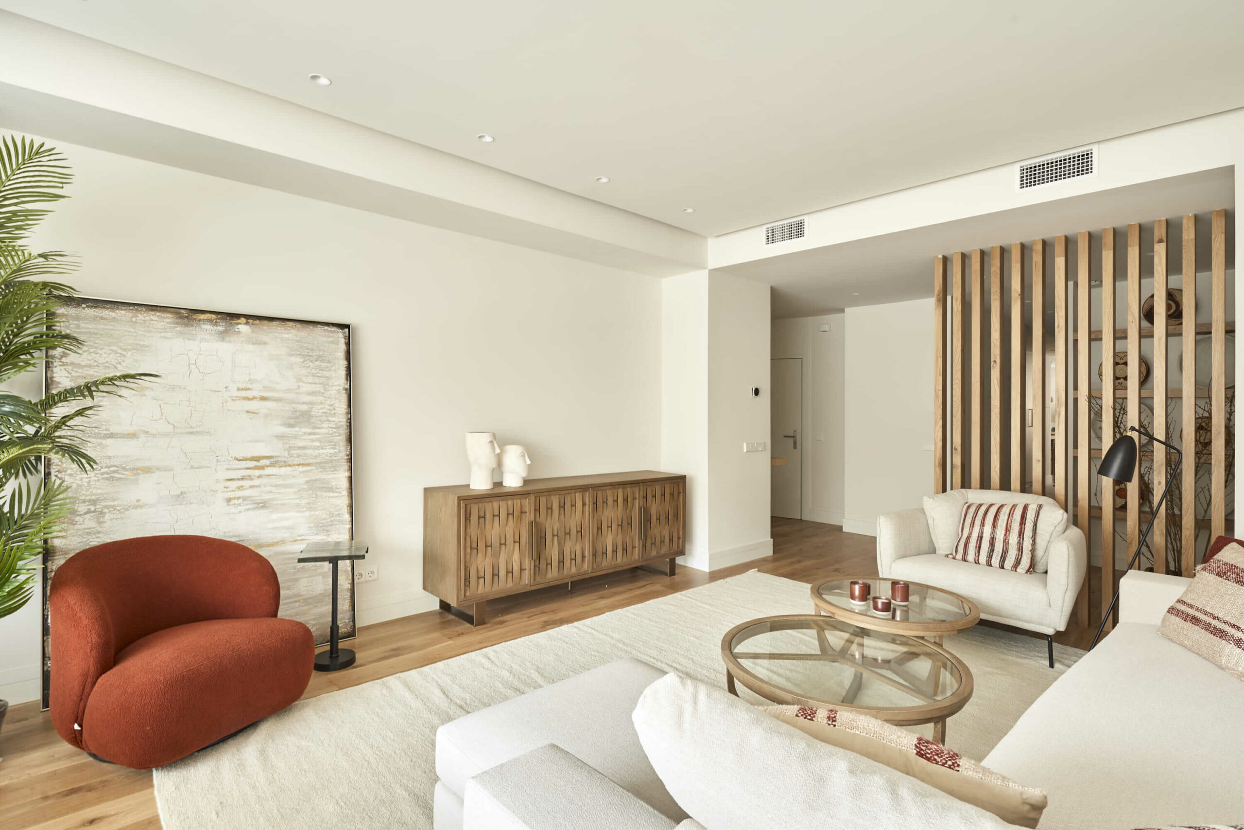 Salón contemporáneo con butaca roja, sofá claro, mesa de centro de cristal, aparador de madera y panel decorativo de listones verticales.