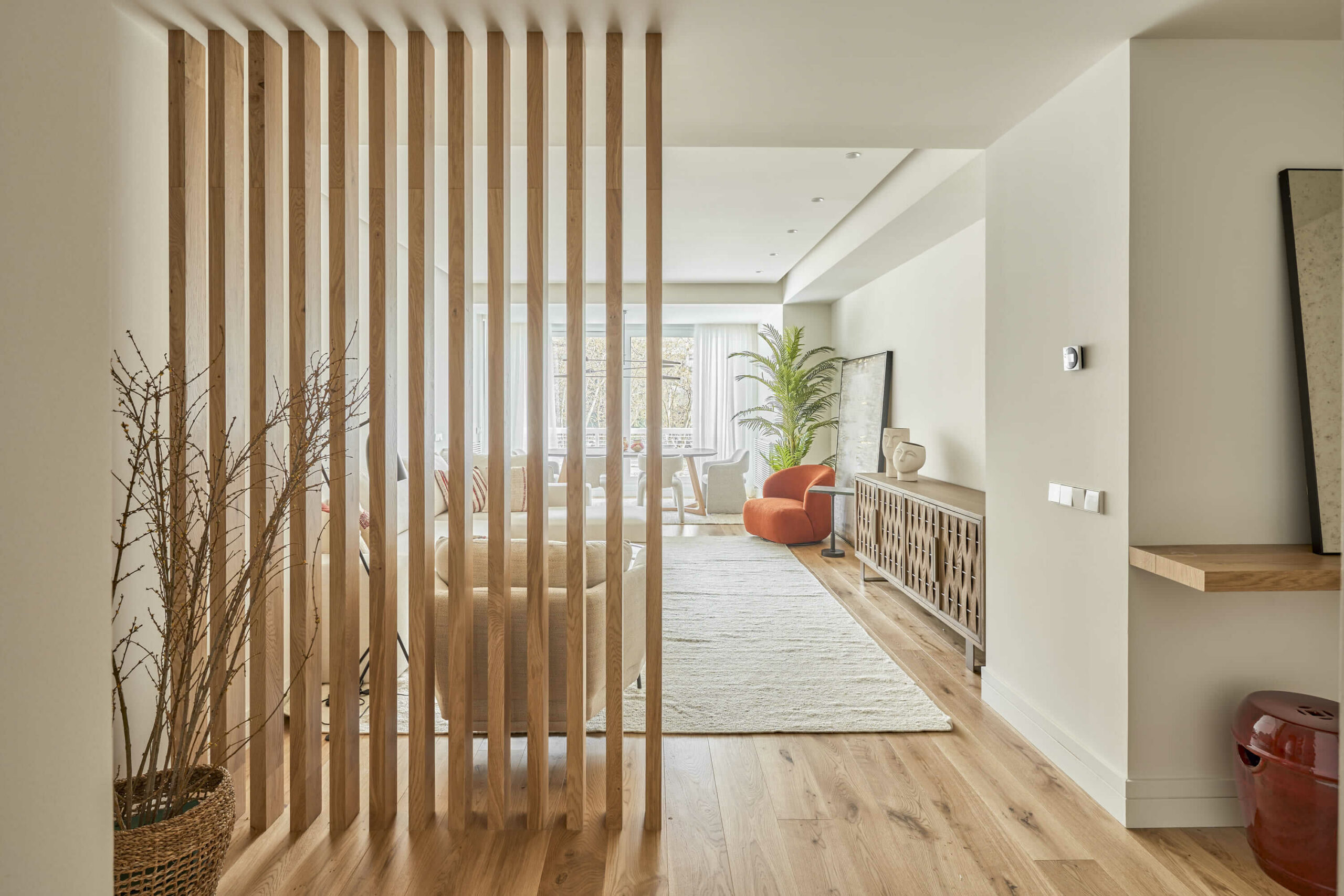 Entrada con panel de listones de madera que divide visualmente el salón, donde se aprecian suelos de madera, butaca roja y decoración en tonos neutros.