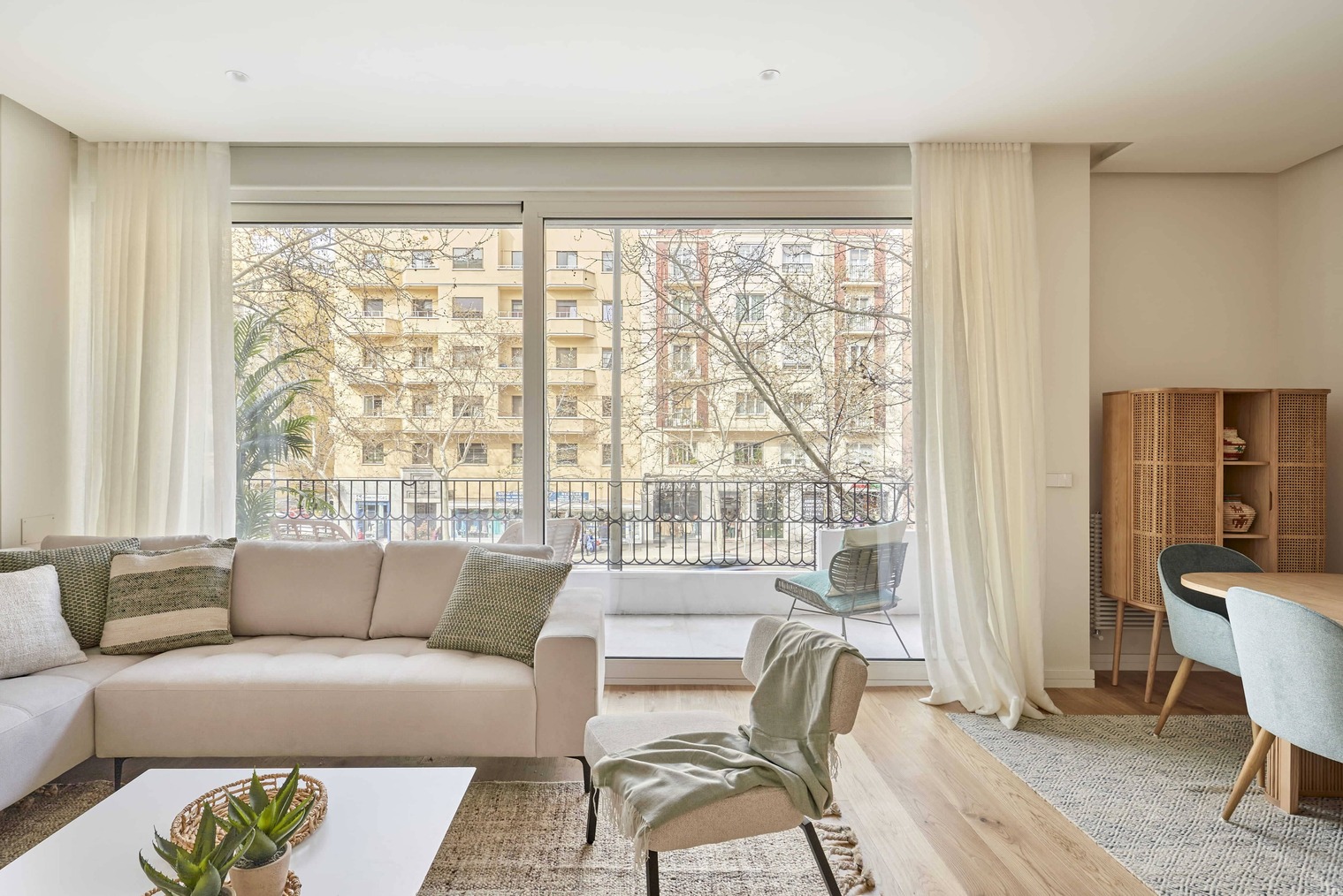 Salón luminoso con gran ventanal, sofá en tonos neutros, sillón ligero y comedor con sillas azules, con salida a una terraza exterior.