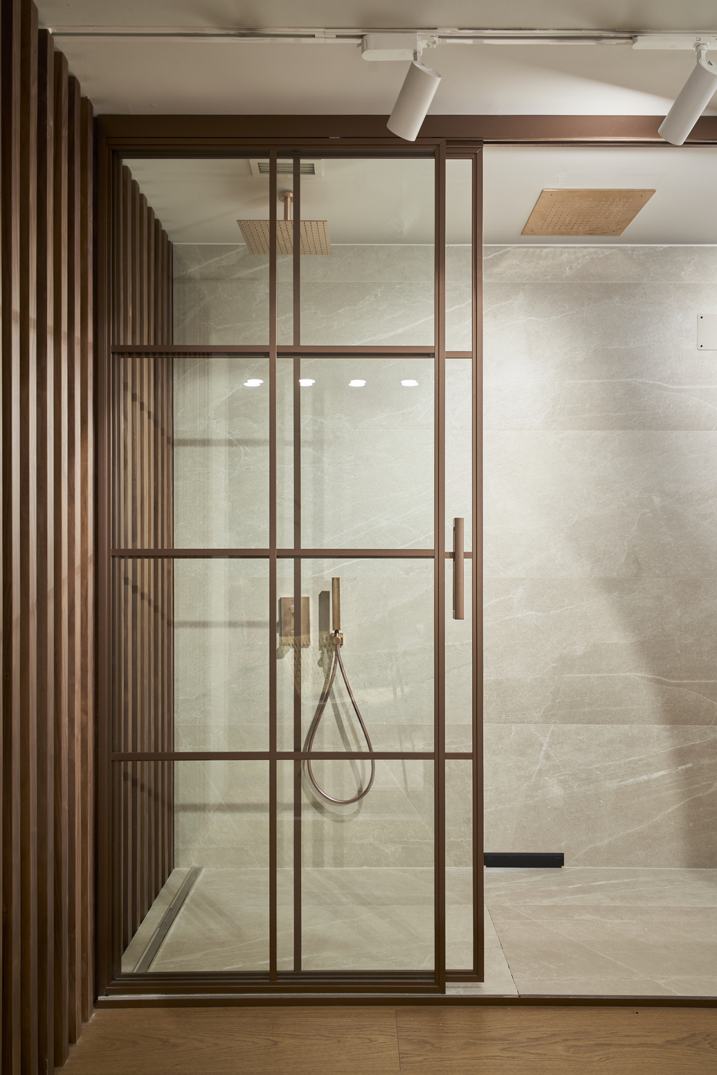 Ducha contemporánea con mampara de vidrio y perfiles metálicos en tono cobre, junto a paredes de mármol claro.