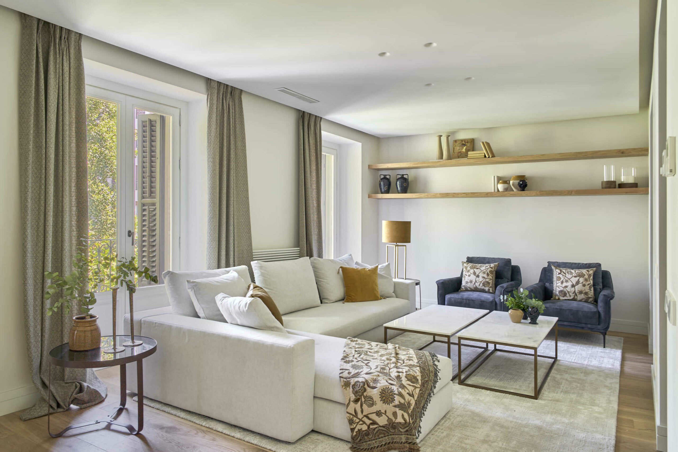 Salón luminoso con sofá claro y estantería de madera integrada Salón luminoso con sofá modular en tonos claros, dos butacas azules, mesa de centro doble con estructura metálica y estantería de madera integrada en la pared.