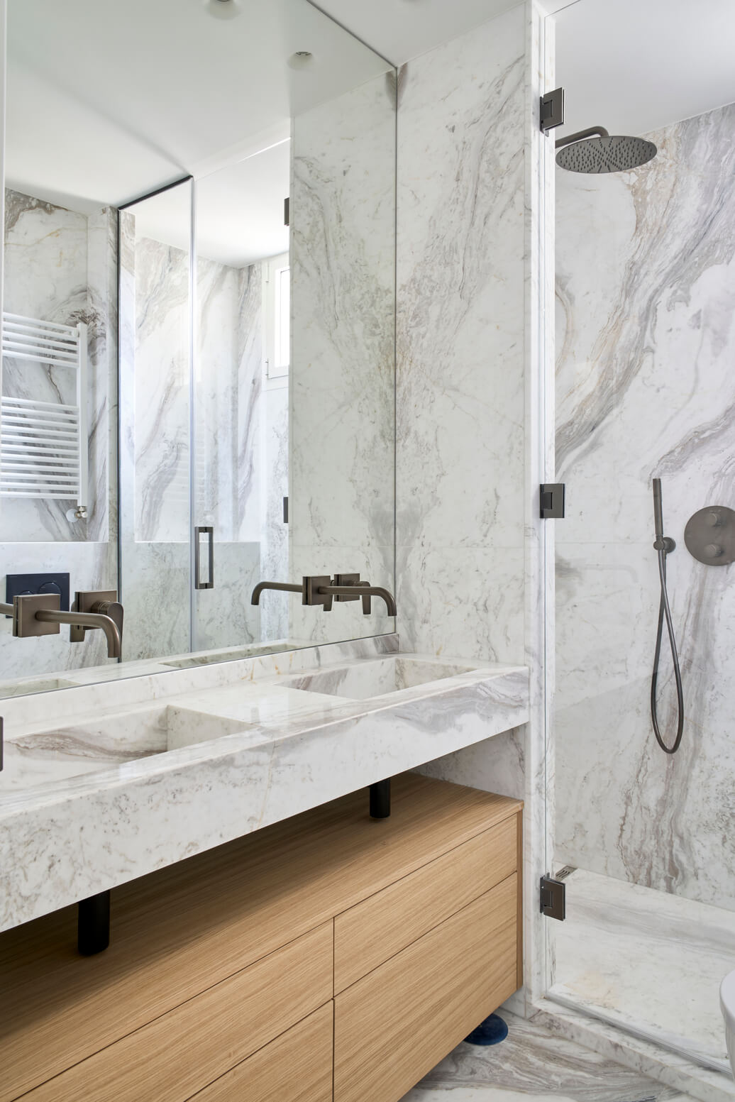 Baño moderno con lavabo doble en mármol vetado, grifería oscura y ducha con mampara de vidrio.