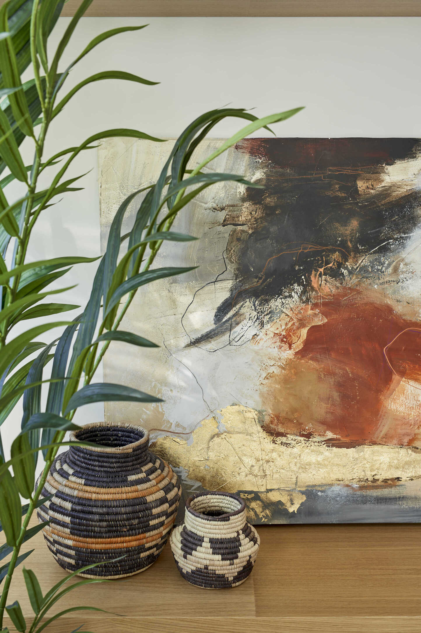 Composición decorativa de estilo vintage con cestas artesanales de fibras naturales, planta verde y cuadro abstracto en tonos terrosos y dorados.