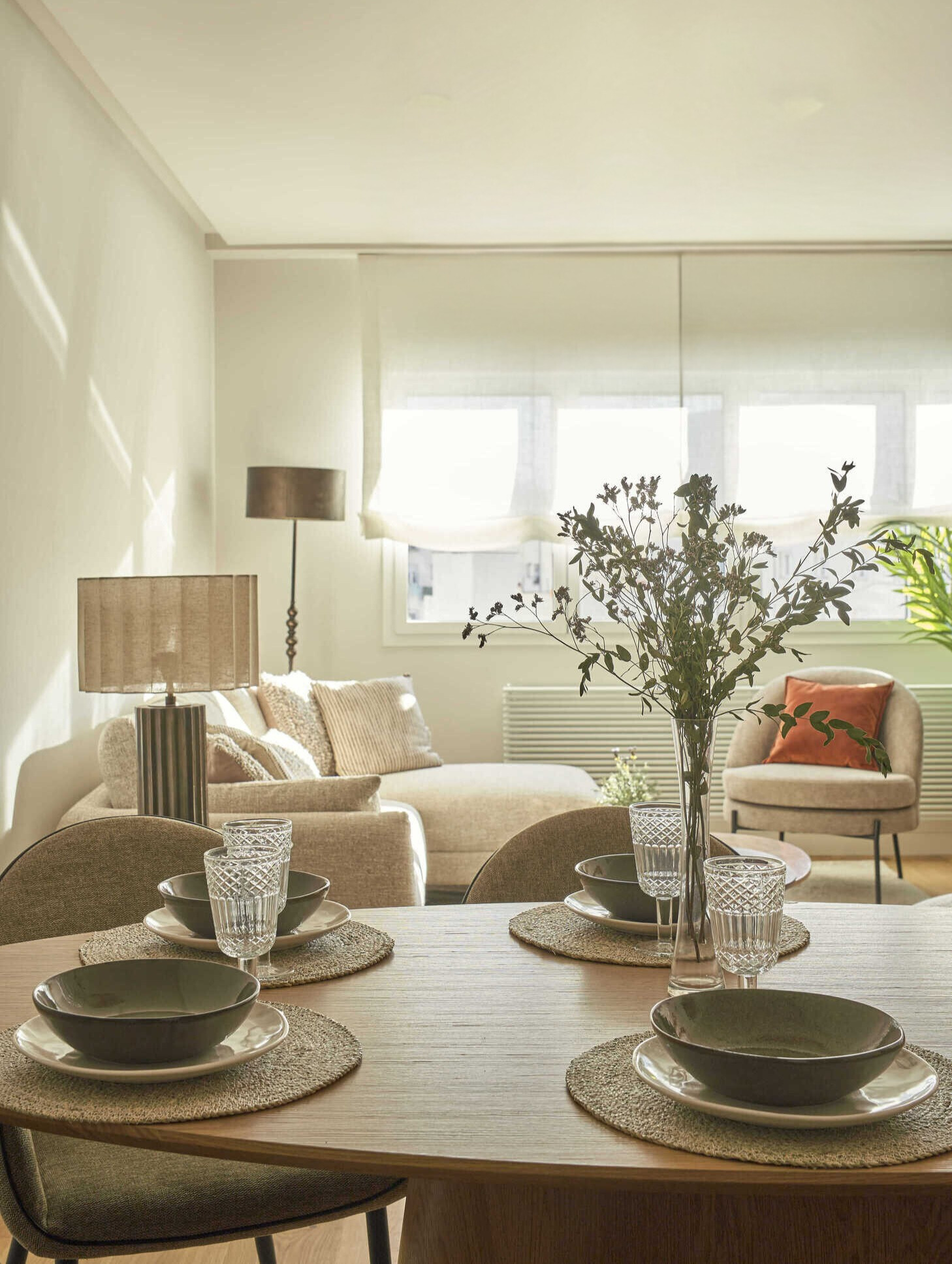 Comedor con mesa puesta y vajilla neutra, junto a un salón luminoso con sofá claro, lámparas decorativas y butaca con cojín terracota.