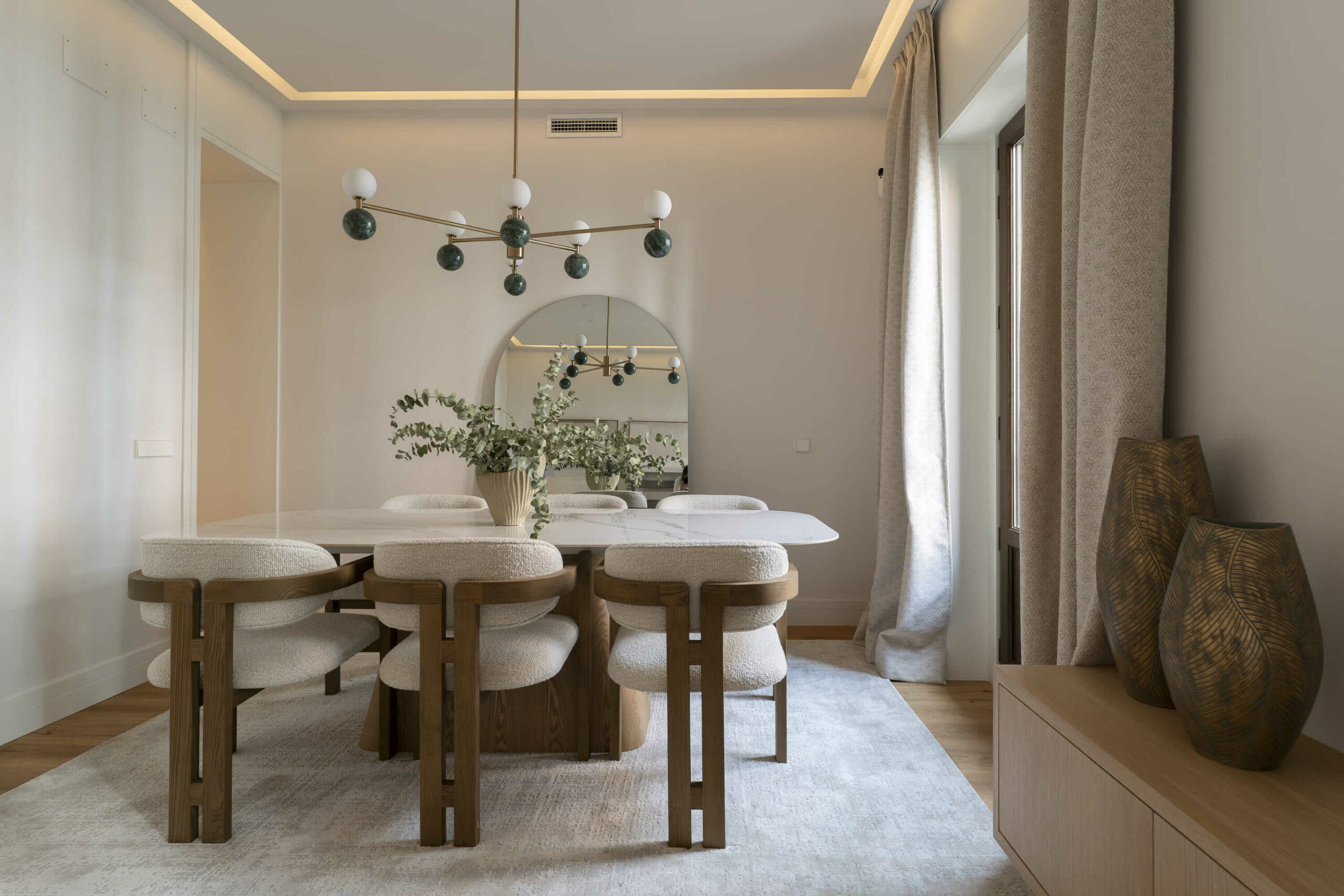 Comedor sofisticado con mesa de mármol, sillas tapizadas en bouclé, lámpara escultórica y gran espejo arqueado que amplifica la luz natural.