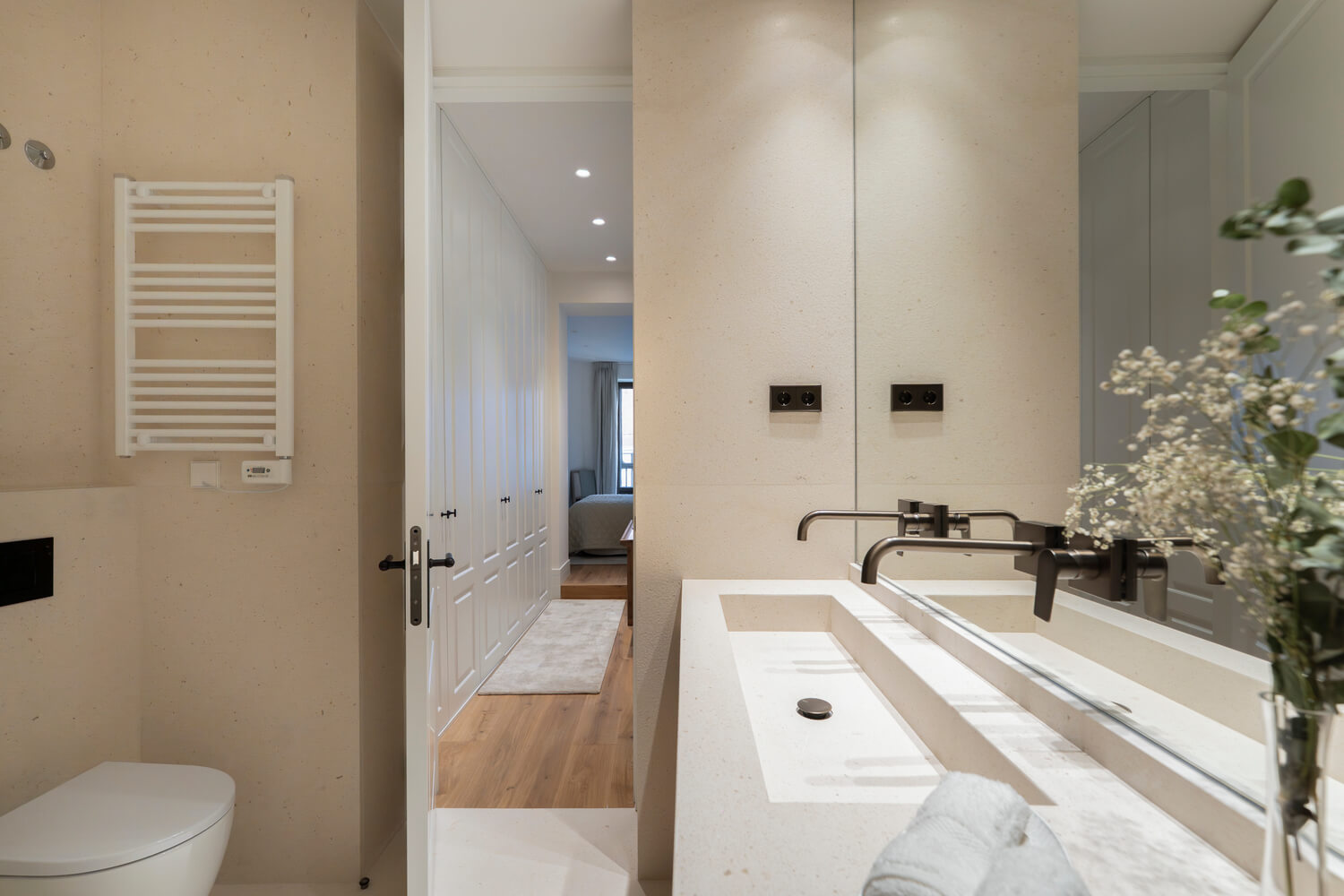 Baño moderno en tonos claros con lavabo integrado, grifería negra y vista hacia el dormitorio a través de una puerta abierta.