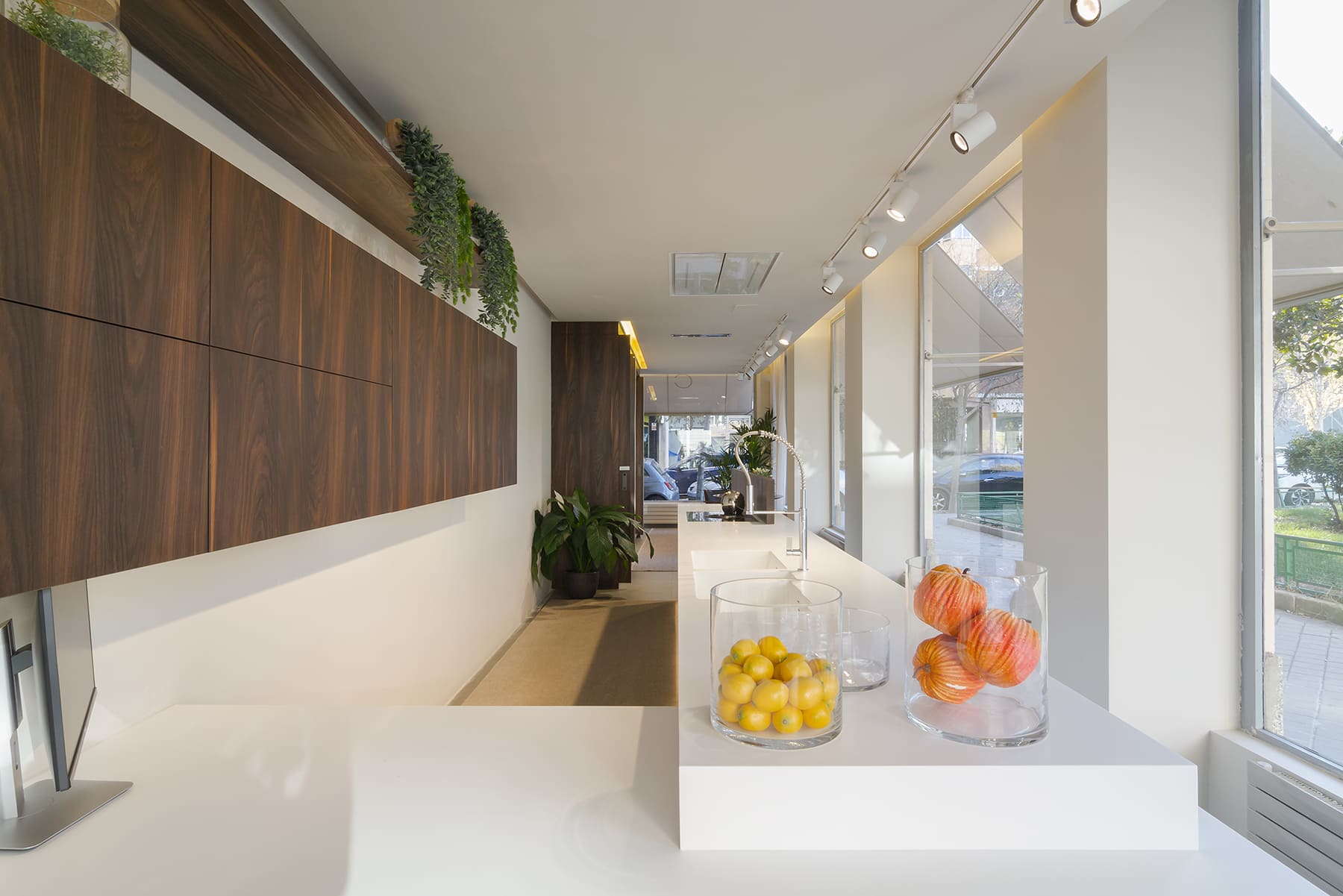 Showroom de exposición de cocinas con encimeras blancas, módulos altos en madera oscura y elementos decorativos como frutas en tarros de cristal.